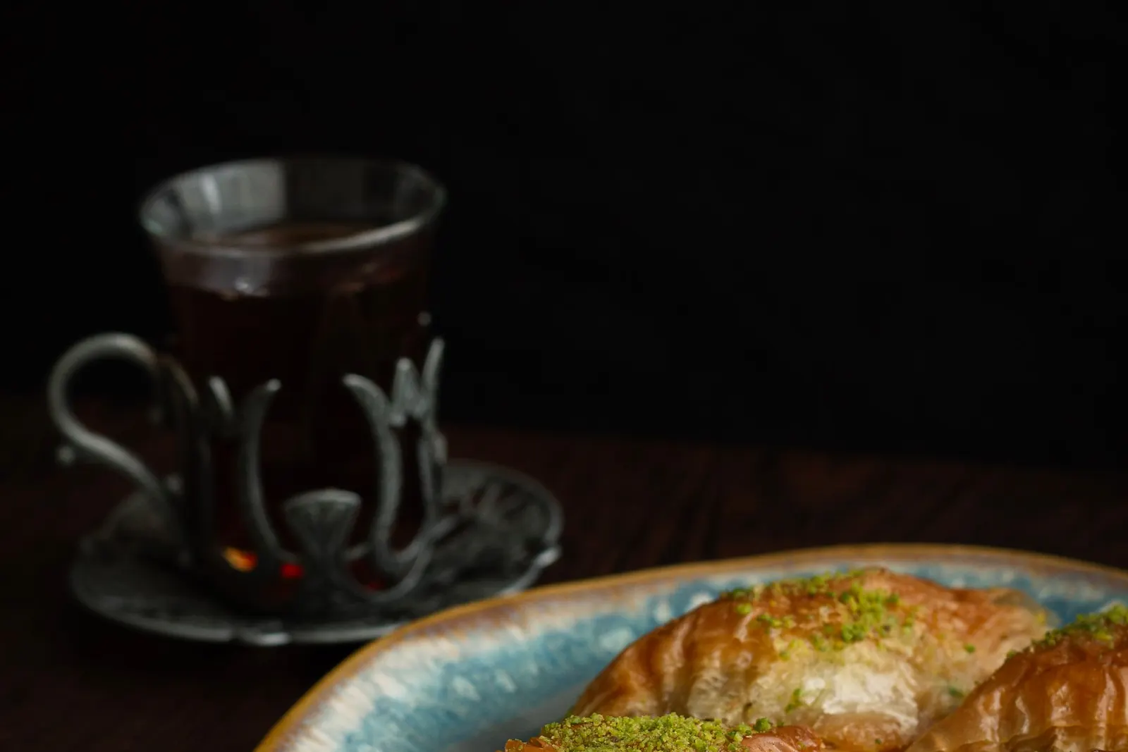 Pistachio baklava on a ceramic plate beside a glass of hot tea in a metal holder, on a dark table