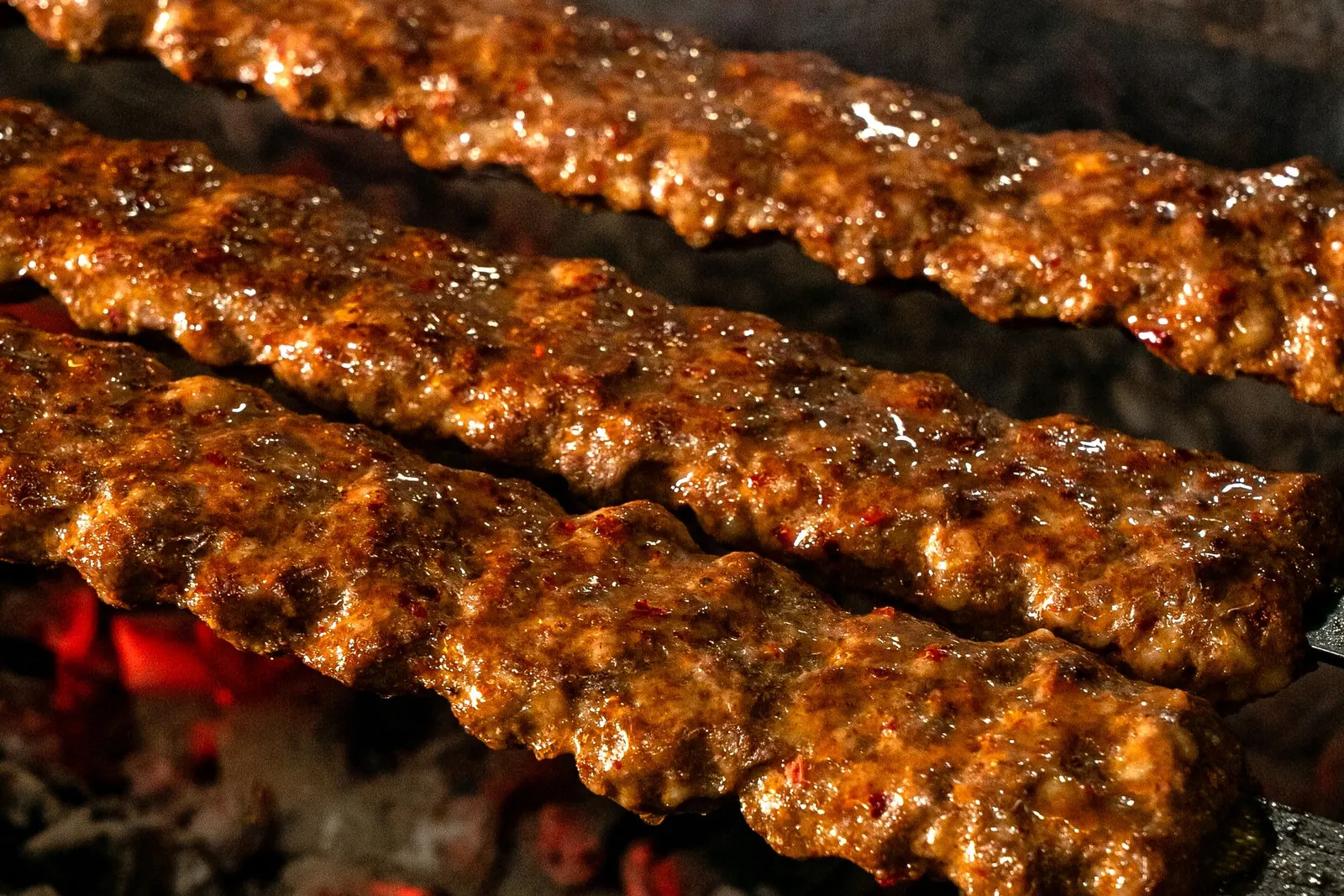Kebabs on flat skewers grilling over glowing hardwood coals, smoke rising through the dark